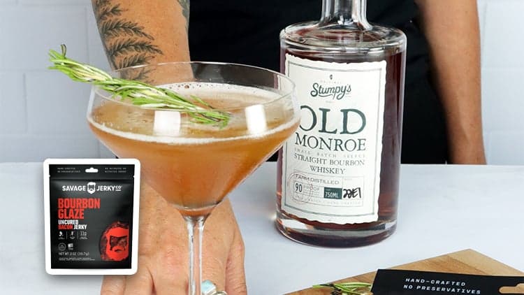 A glass of Smoked Rosemary Bourbon Sour being offered by a hand in front of a bottle of Sumpy's Old Monroe Bourbon Whiskey.