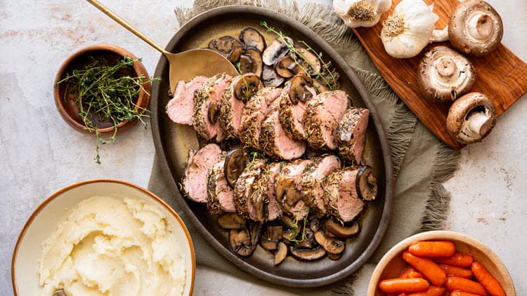 Roasted Garlic Mushroom Pork Tenderloin shown served on a plate with bowls of mashed potatoes and carrots sitting nearby.
