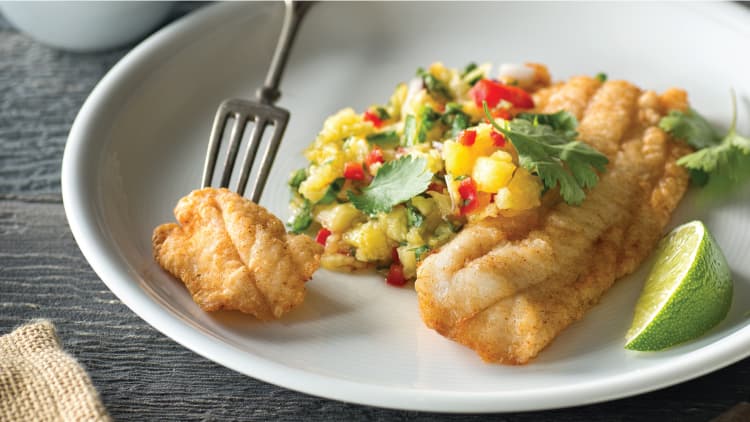 A fork picking up a piece of Cajun Tilapia served on a plate with Pinapple Salsa.