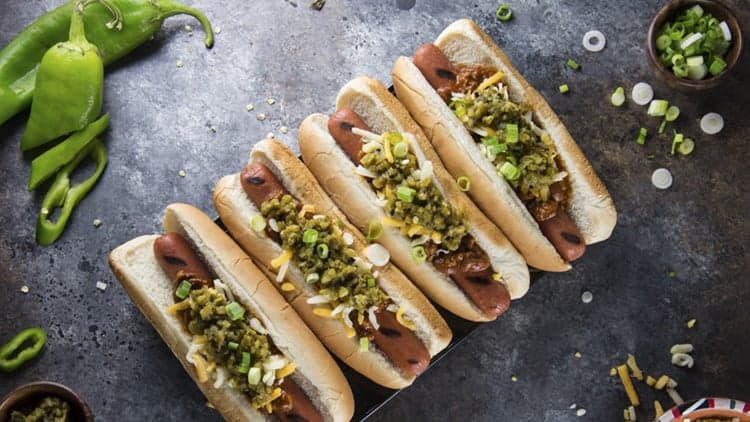 A top down view of four Hatch Chile Hotdogs on a board surrounded by the ingredients used to make them.