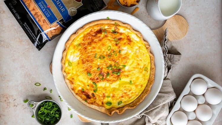Three Cheese Quiche shown cooked and ready to serve, next to eggs and a bag of Dierbergs Cheddar Cheese.